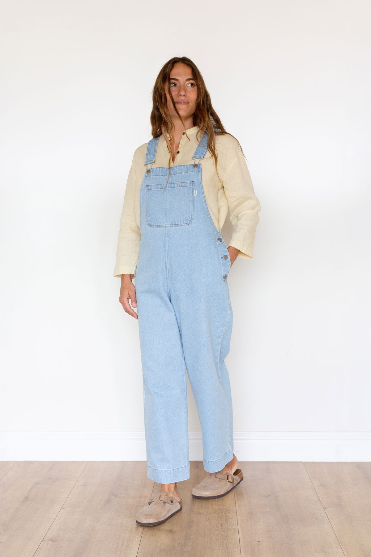 Woman wearing light blue denim overalls and a yellow linen shirt with one hand in pocket.