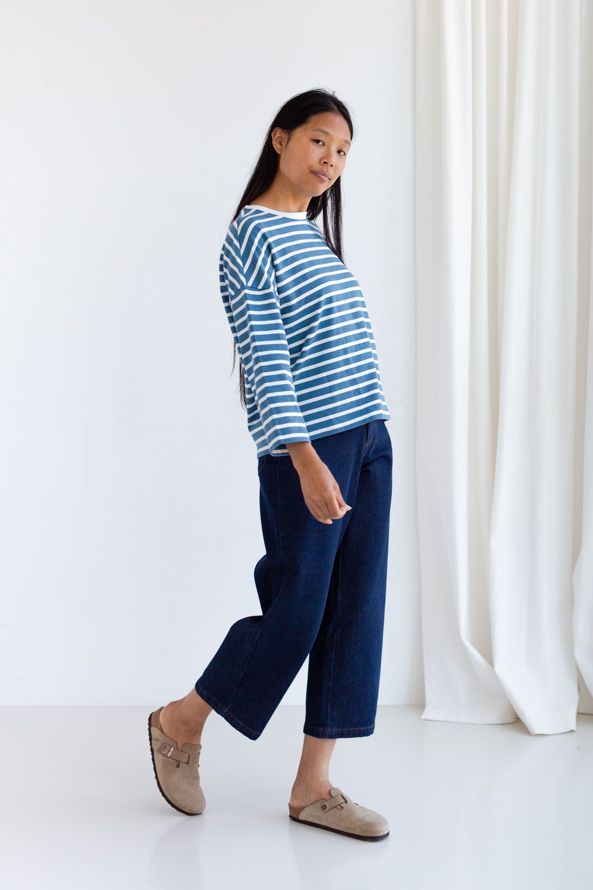 Full body view of woman wearing a dusty blue and white striped sweater and dark blue jeans.