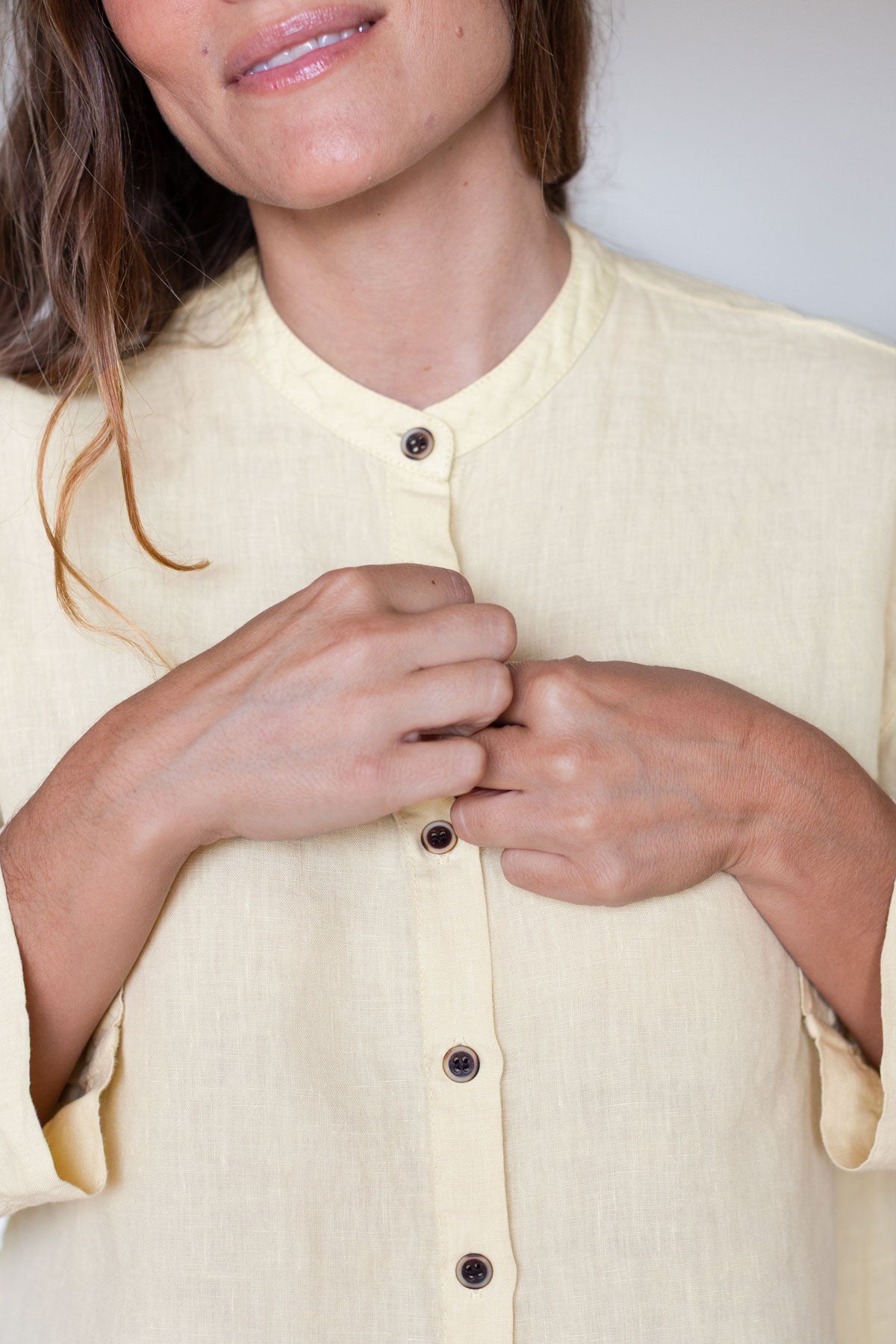 Close-up of a woman unbuttoning a gardenia yellow linen shirt with a mandarin collar.