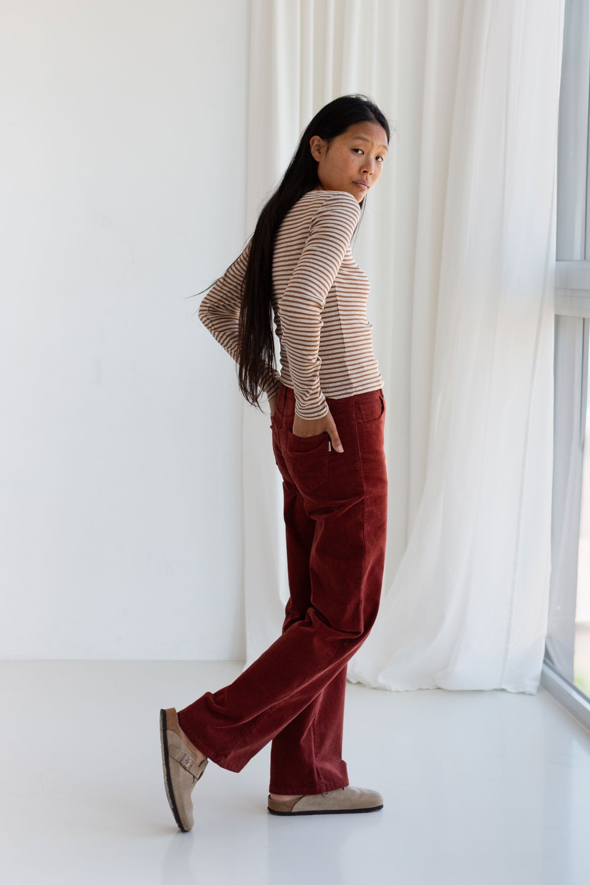 Full body back view of a woman walking in a caramel striped top and red corduroy pants.