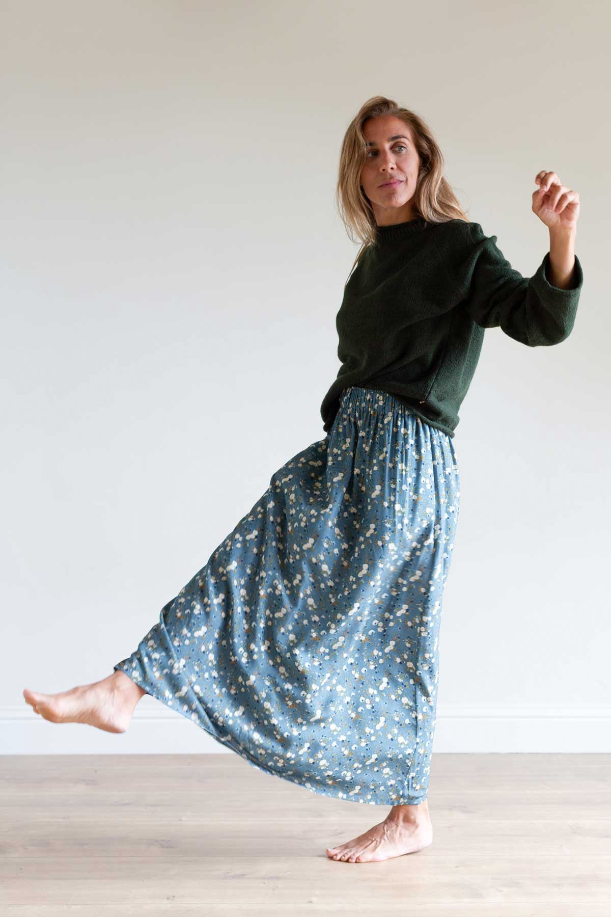 Full body shot of a woman playfully lifting her leg in a blue floral muslin maxi skirt.